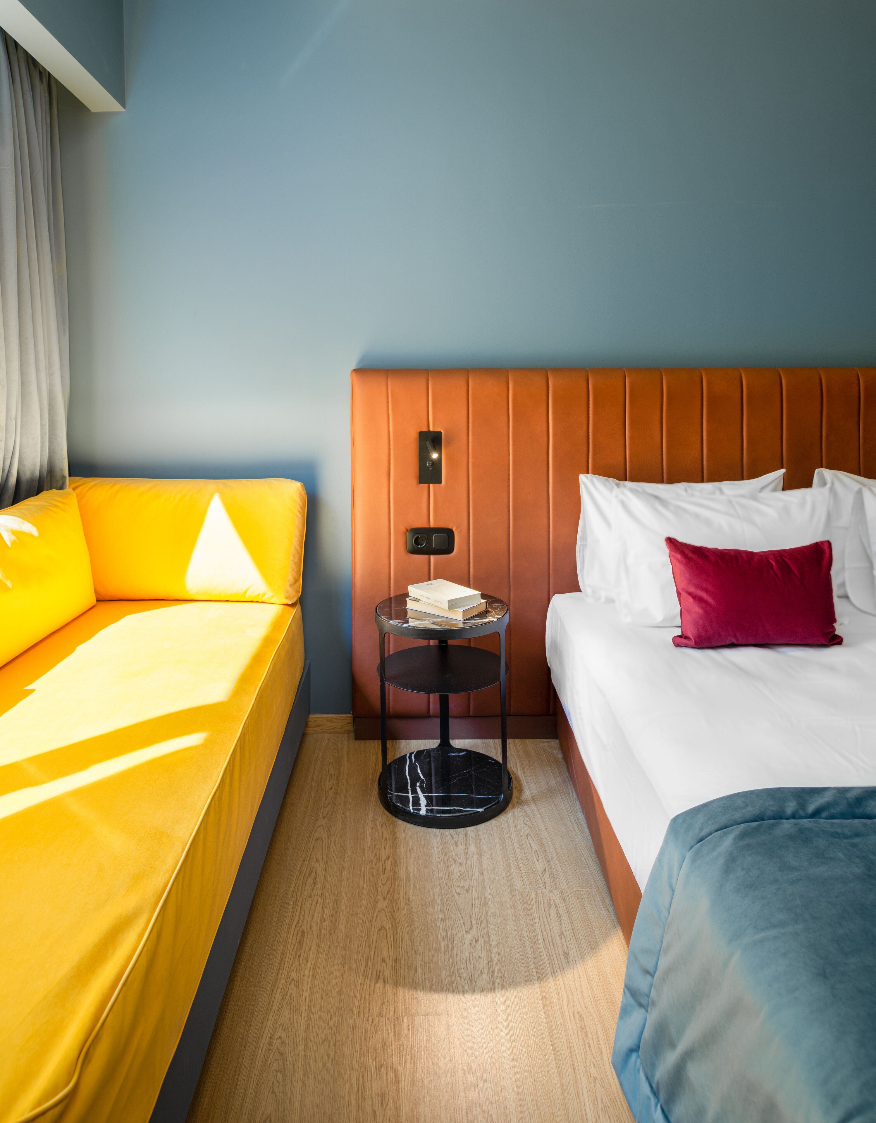 Ein Hotelzimmer mit gelbem Sofa, einem Bett mit rotem Kissen, einem kleinen runden Tisch mit einem Buch und einem hölzernen Kopfteil.