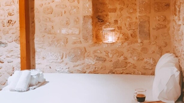 Une pièce avec un mur en pierre, une poutre en bois, un lit recouvert de draps blancs, une petite table avec une tasse et un livre, et une fenêtre avec une cage à oiseaux.