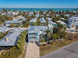 A coastal residential area with houses featuring modern architecture, a prominent house with a large balcony and glass doors, and a view of the ocean and a bridge in the distance. - Still Waters Beach House- A north end tropical getaway. (Anna Maria)