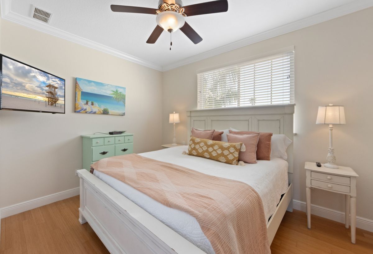 A bedroom with a bed, a ceiling fan, a nightstand, a lamp, and two paintings on the wall.