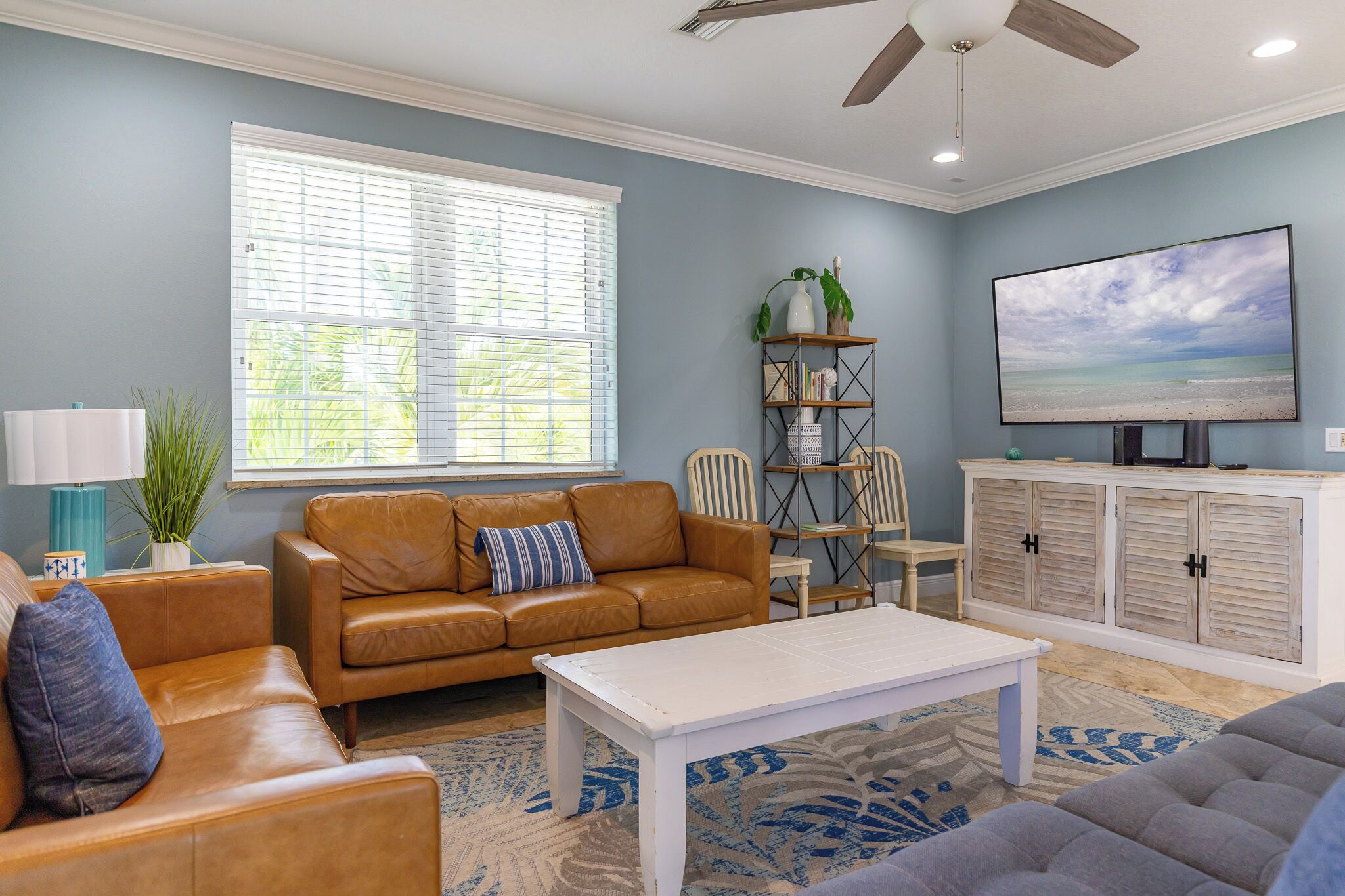 A living room with a brown leather sofa, a white coffee table, a blue patterned rug, a bookshelf, and a television on a white cabinet.