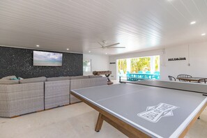 A modern living room with a ping pong table, a large sofa, a flat-screen TV, and a ceiling fan.