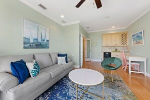 A living room with a sofa, a round coffee table, and a green chair. There is a kitchen in the background with cabinets and a dining table. - Still Waters Beach House- A north end tropical getaway. (Anna Maria)