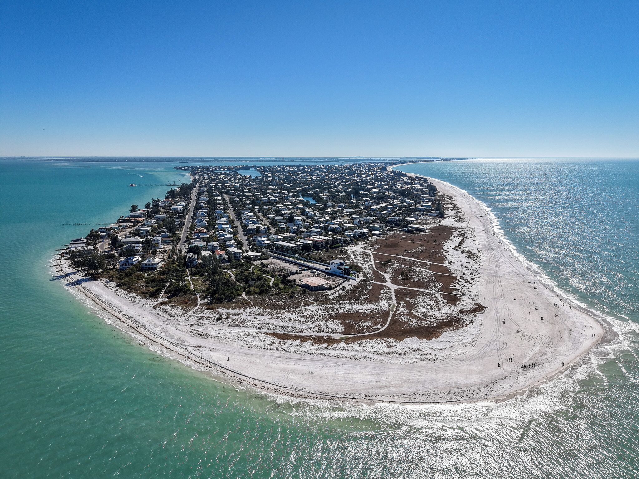 An aerial view of a coastal area with a densely packed residential zone, a sandy beach, and clear blue waters.