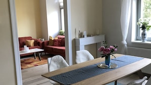 Un comedor con una mesa de madera, sillas blancas y un mantel individual azul. Hay una ventana con cortinas blancas y un florero con flores sobre la mesa.