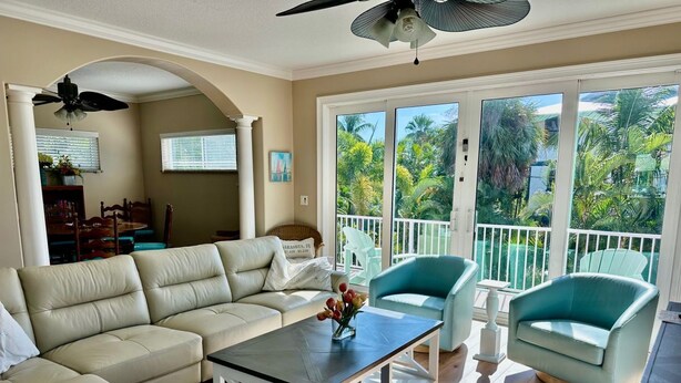 A spacious living room with a beige sectional sofa, a coffee table, and teal armchairs. A ceiling fan is visible, and the room has large windows with a view of palm trees.