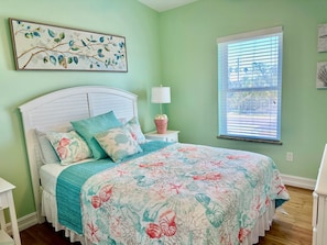 Quarto com cama, mesa de cabeceira, abajur, janela com persianas e quadro na parede.