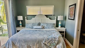 A bedroom with a large bed, two bedside tables with lamps, a window with blinds, and a framed picture on the wall.