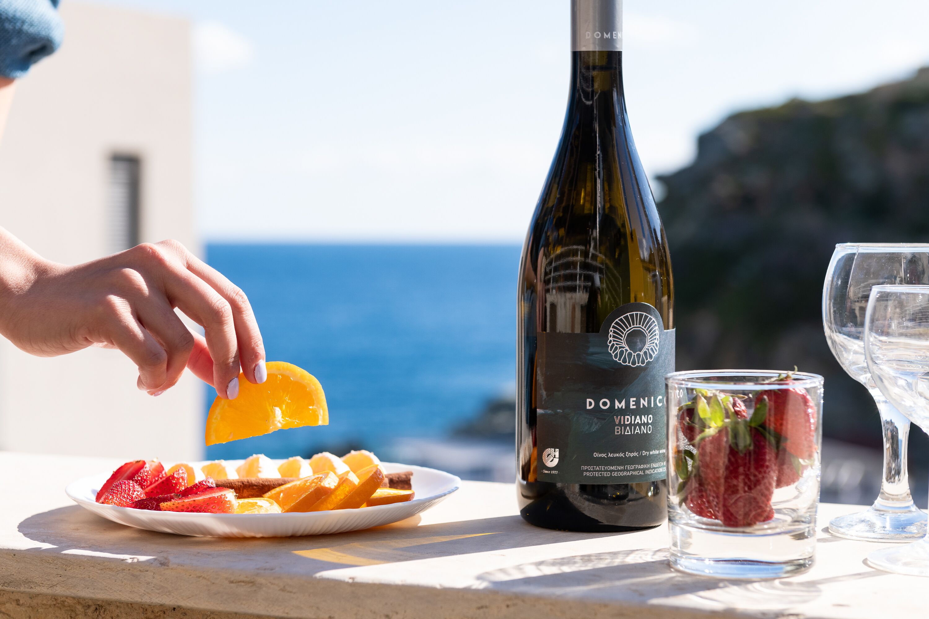 A bottle of wine, a glass of water, and a plate of fruit on a table with a scenic view.