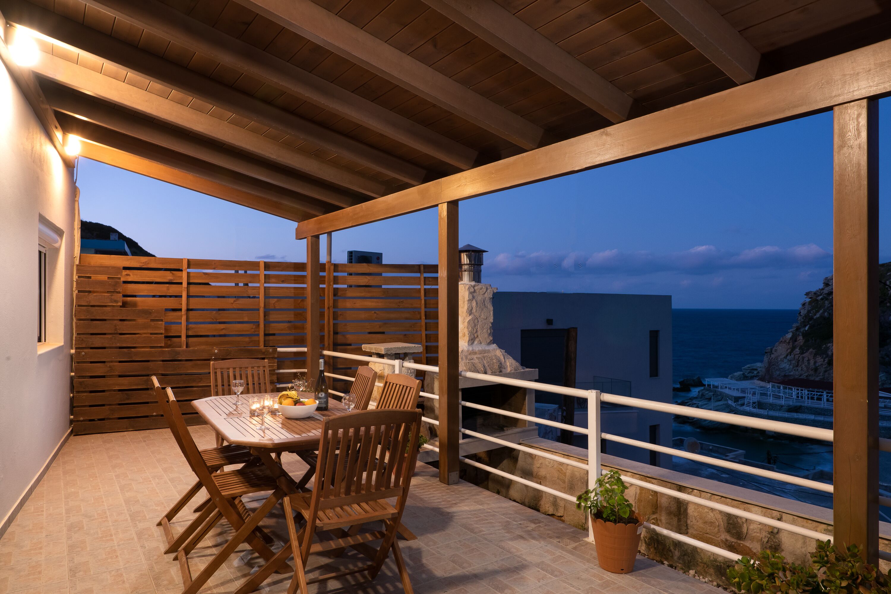 A wooden deck with a table and chairs, overlooking a coastal area with a rocky landscape and a building in the distance.