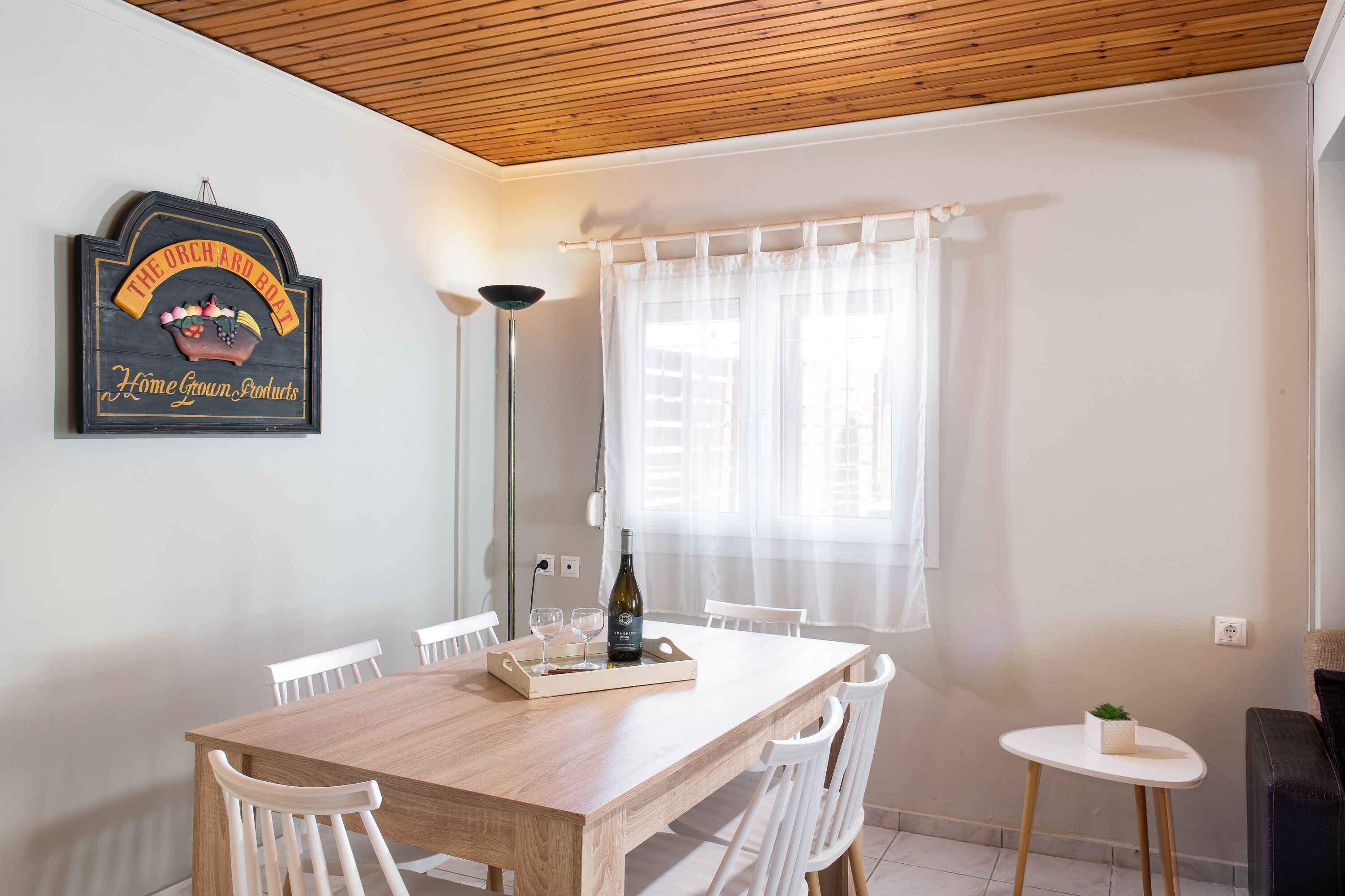 Zona de menjador amb taula de fusta, cadires blanques, taula auxiliar petita i un cartell penjat a la paret.