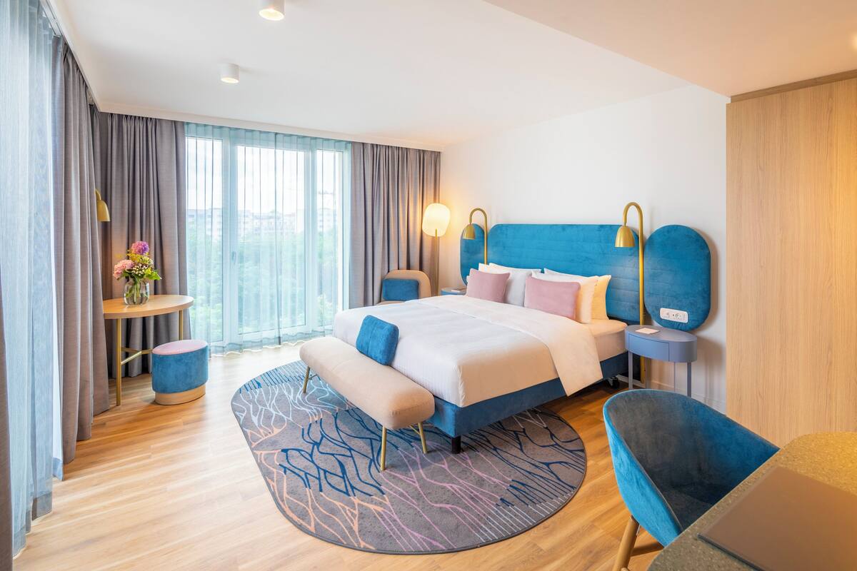 A modern hotel room with a large bed, a round rug, a blue armchair, and a small side table with a lamp.