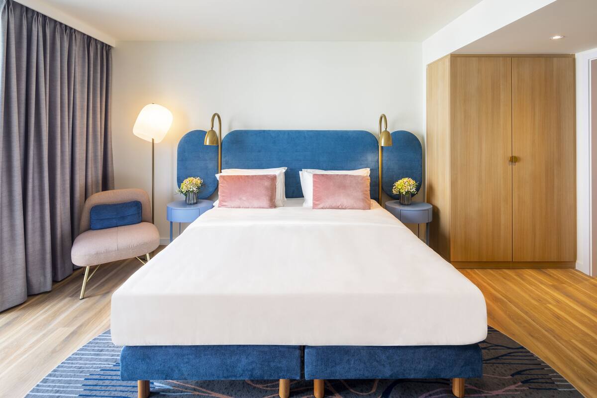 A modern hotel room with a large bed, a blue headboard, a wooden wardrobe, a nightstand with a lamp, and a chair.