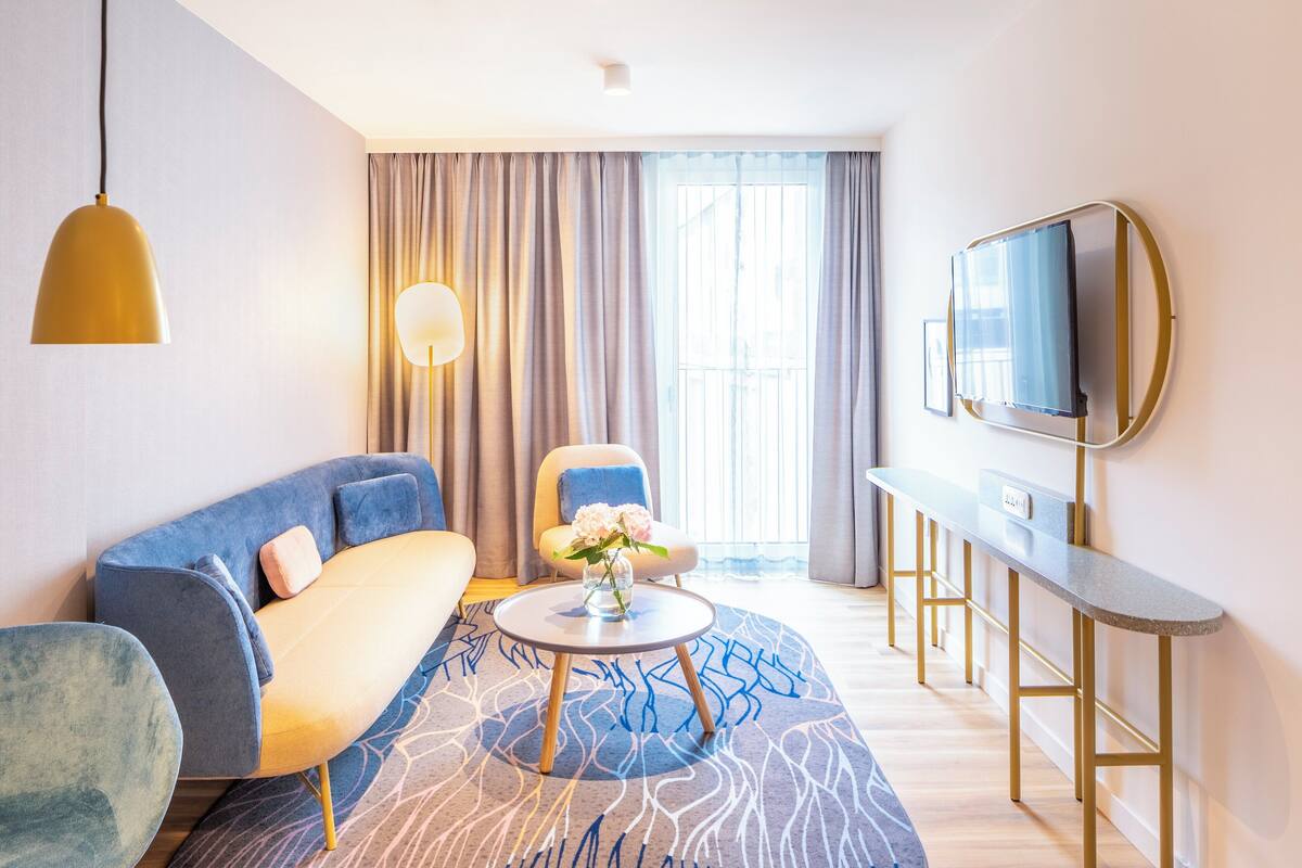 A modern hotel room with a sofa, armchair, round coffee table, and a TV mounted on the wall.