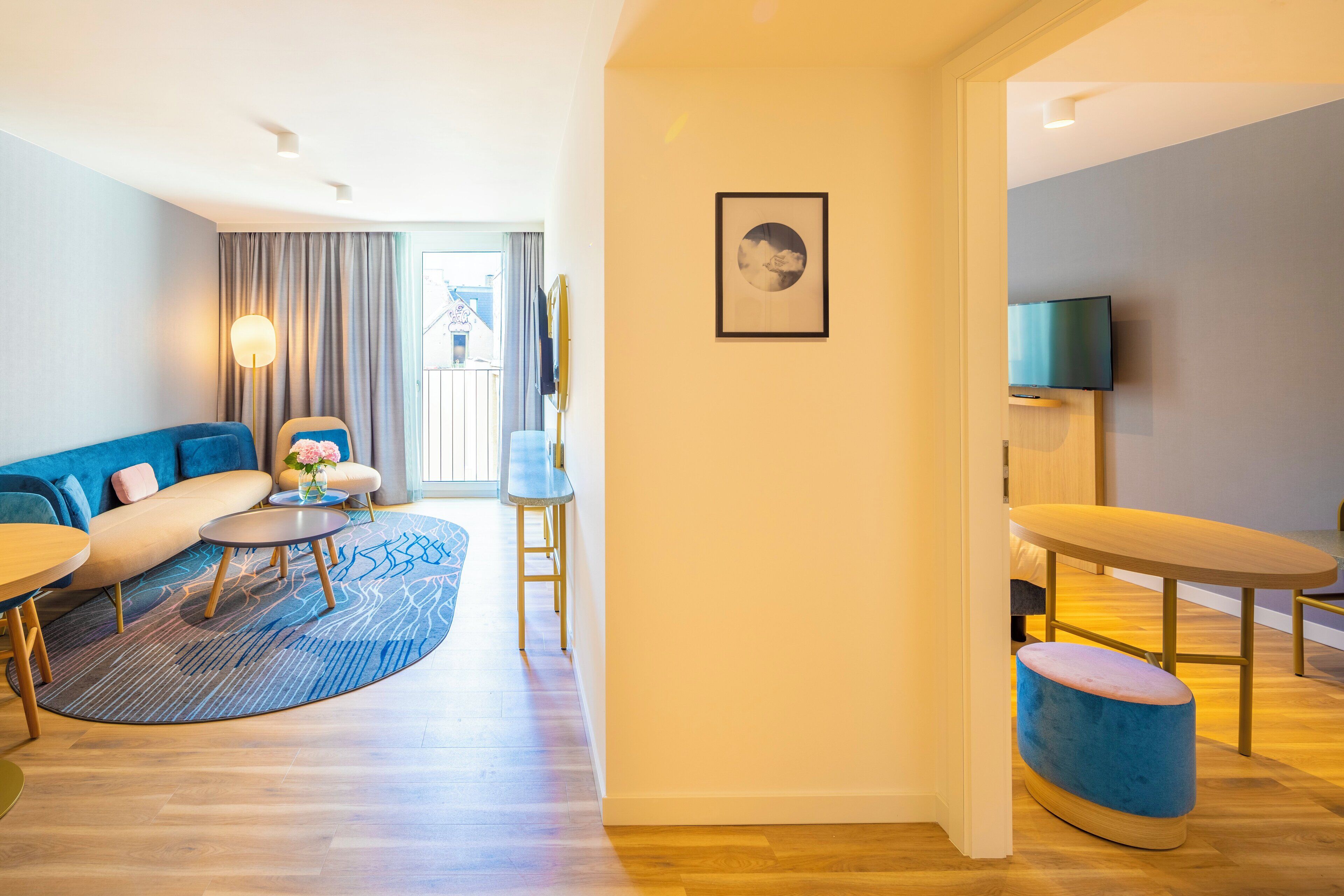 Quarto de hotel moderno com sofá, duas mesas redondas, um pequeno banquinho, televisão e um quadro na parede.