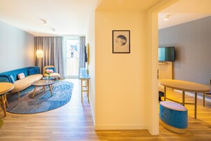 A modern hotel room with a sofa, two round tables, a small stool, a TV, and a framed picture on the wall.