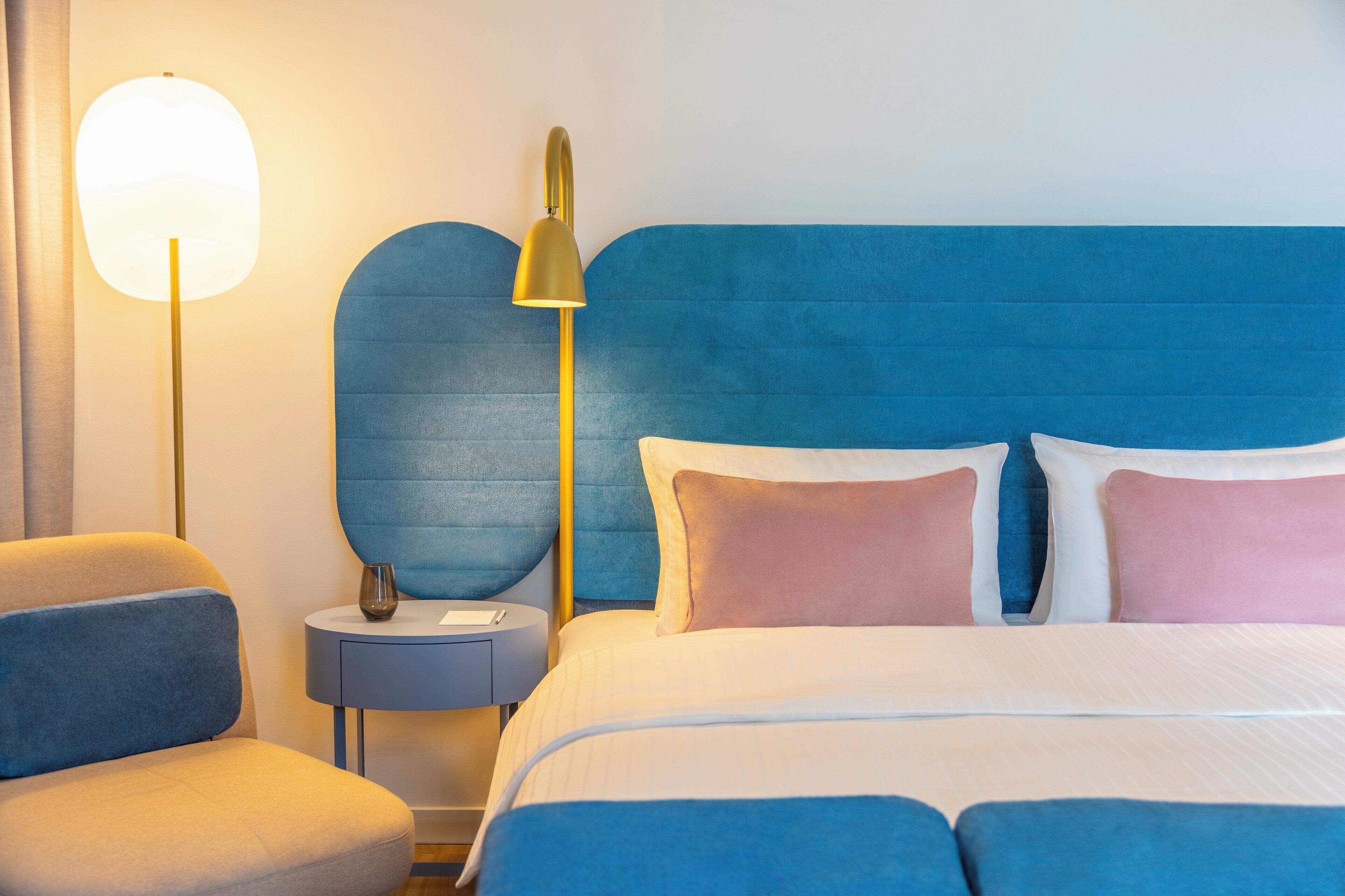 Una habitación de hotel moderna con un cabecero tapizado en azul, una mesita de noche con lámpara, y una cama con ropa de cama blanca y rosa.