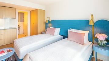 A hotel room with two beds, a small table, a nightstand with a lamp, and a view of the kitchen.