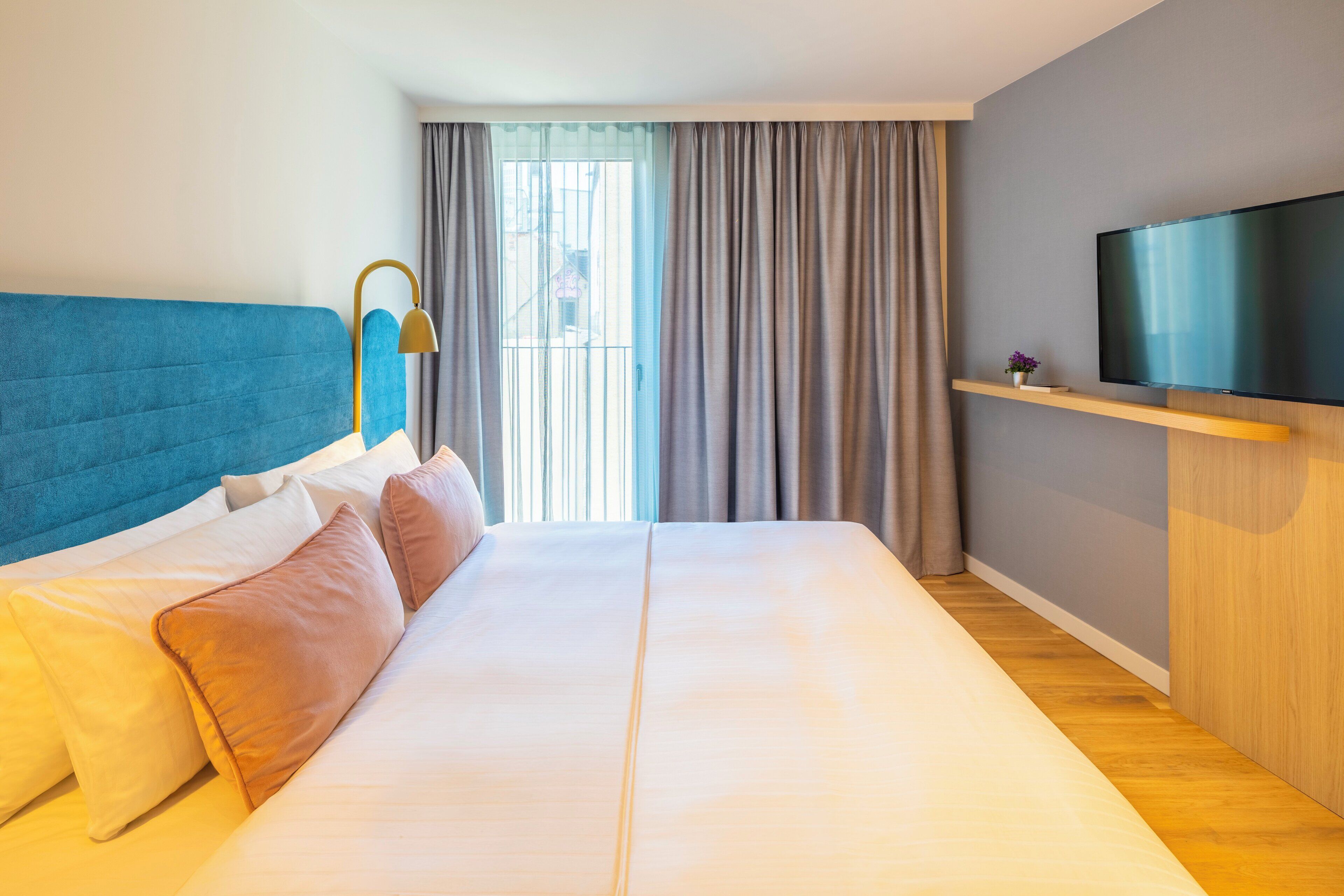 A hotel room with a large bed, a TV mounted on the wall, and a window with curtains.