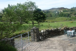 Un paysage rural avec un mur de pierres, un portail en métal et une table entourée de chaises à l’extérieur.