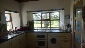 Eine Küche mit Spüle, Herd, Backofen und Waschmaschine. Ein Fenster bietet Blick auf Bäume, und an der Wand hängt eine Uhr.