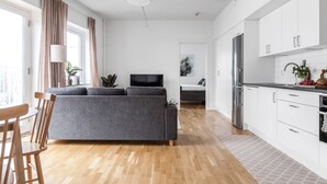 Una sala moderna con un sofá gris, una mesa de comedor de madera y sillas. Se ve una cocina con gabinetes blancos y un refrigerador de acero inoxidable.