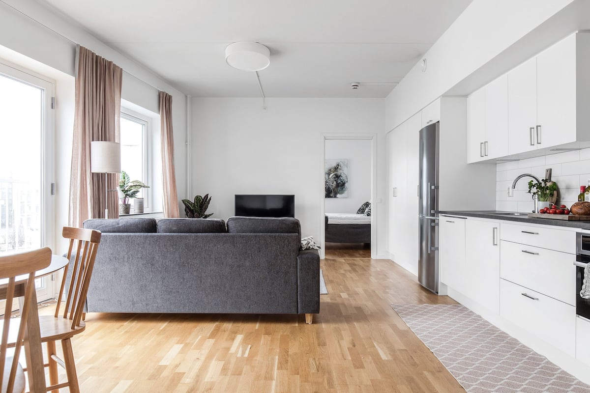Ein modernes Wohnzimmer mit einem grauen Sofa, einem Holzespeisetisch und Stühlen. Man sieht eine Küche mit weißen Schränken und einem Edelstahl-Kühlschrank.