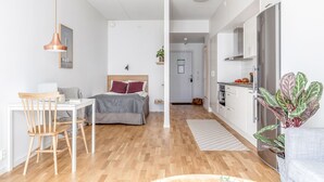 Una habitación moderna y bien iluminada con suelo de madera, una cama con cabecero gris, un escritorio con silla, una lámpara colgante y una zona de cocina con refrigerador y planta.