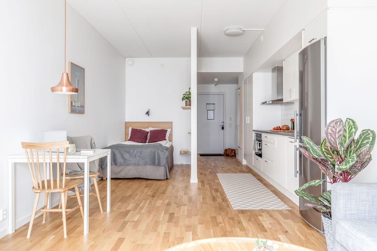 Ein modernes, gut beleachtetes Zimmer mit Holzboden, einem Bett mit grauem Kopfteil, einem Schreibtisch mit Stuhl, einer hängenden Pendelleuchte und einer Küchenzeile mit Kühlschrank und Pflanze.