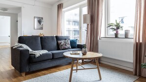 Una sala de estar moderna con un sofá gris, una mesa redonda de madera y vistas a un paisaje urbano a través de grandes ventanales.