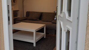 Et værelse med en hvid dør, et rundt hvidt bord, en grå sofa, en gulvlampe og en reol.