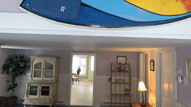 Una habitación con una obra de arte grande y colorida en el techo, un aparador antiguo, una estantería metálica y una lámpara sobre una mesa.