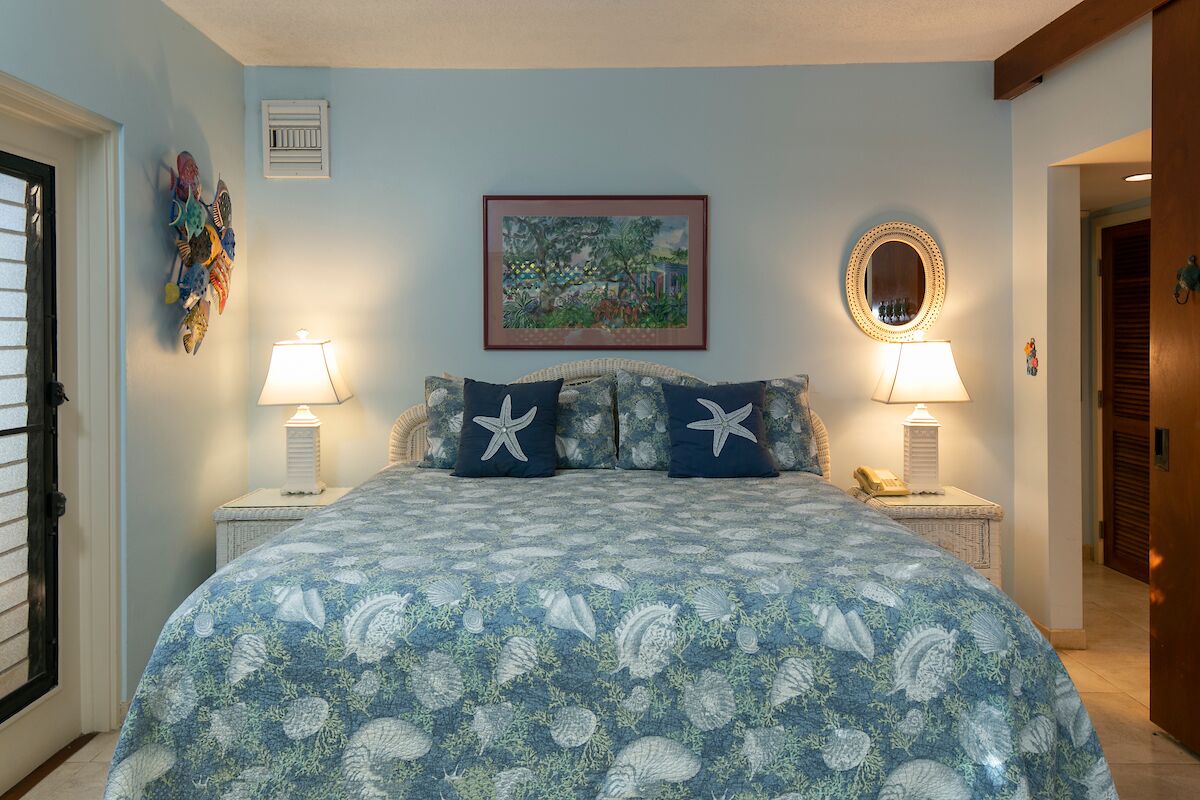 Une chambre à coucher avec un lit recouvert d’une courtepointe à motifs floraux bleus, deux tables de chevet avec des lampes, un tableau encadré au mur et un miroir au-dessus du lit.