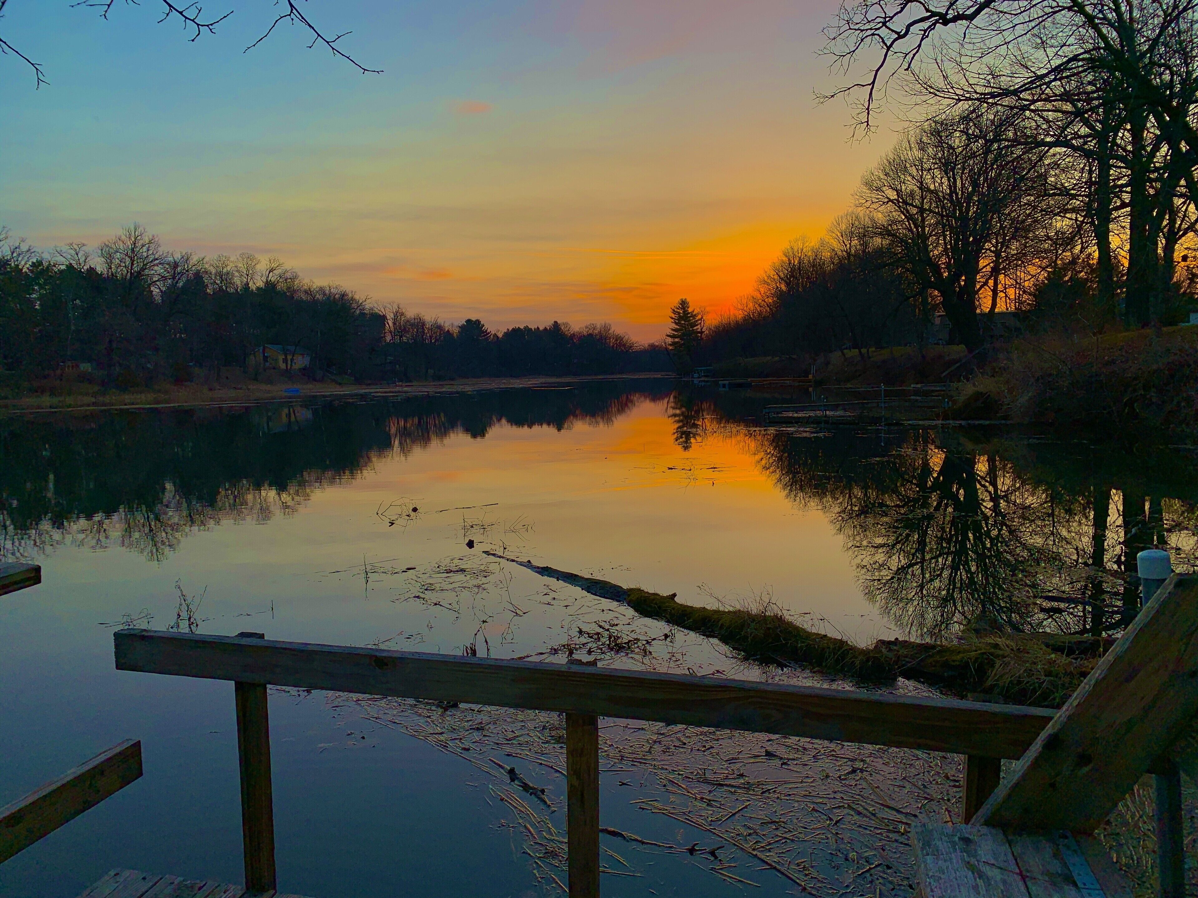 Ein ruhiger Fluss bei Sonnenuntergang mit einem hölzernen Steg und Bäumen im Hintergrund.