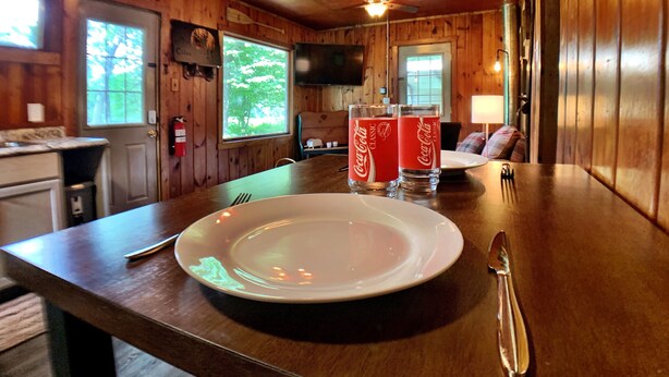 Un interior de madera con una mesa de comedor, dos vasos de Coca-Cola, un plato blanco y un televisor al fondo.