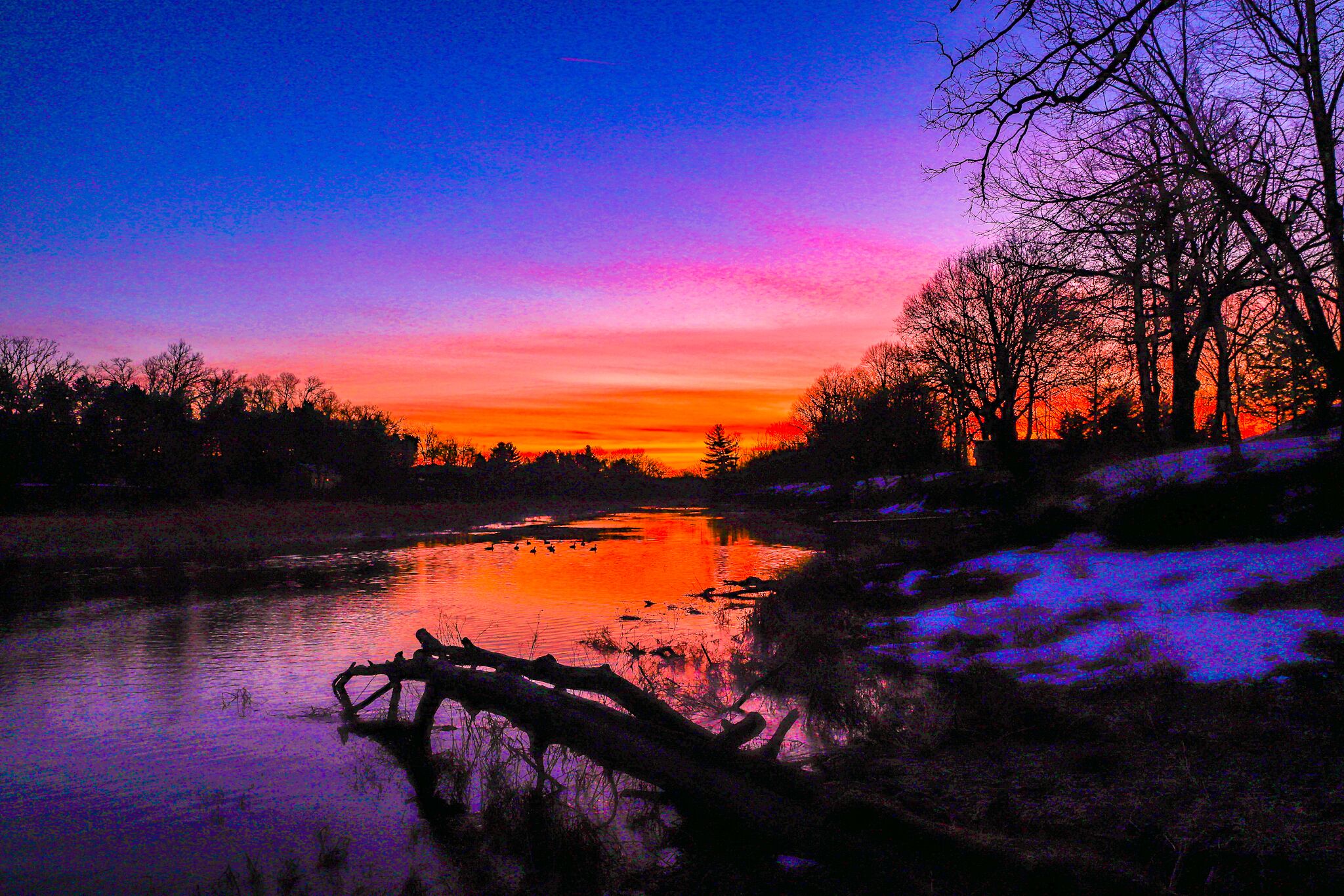 A sunset over a calm river with trees and a snow-covered bank.
