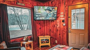 Un interior de cabaña acogedor con televisión, paredes de madera, chimenea y un ventanal grande con vistas a árboles.