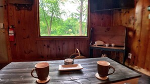 Interior de una cabaña de madera con una mesa dispuesta para dos, una ventana con vistas a árboles y un extintor en la pared.