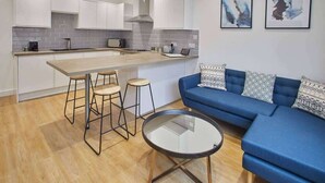 A modern kitchen with white cabinets, a wooden countertop, and a blue sofa with patterned cushions. There are three stools and a glass table. - Host & Stay | Routh Walk (Whitby)