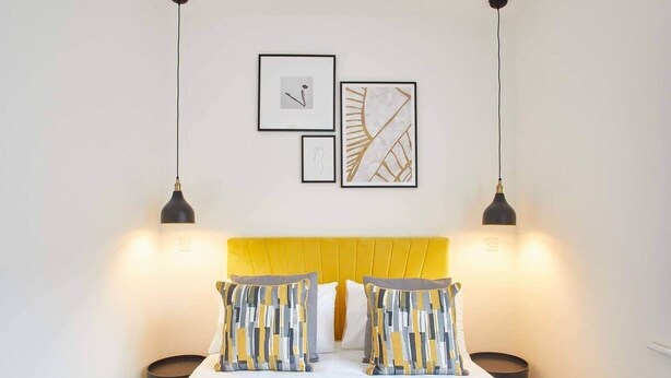 A neatly arranged bedroom with a yellow headboard, two bedside lamps, and three framed artworks on the wall. - Host & Stay | Routh Walk (Whitby)