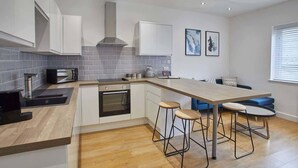 A modern kitchen with white cabinets, a built-in oven, a microwave, and a coffee maker. There are two stools at a wooden table, and a window with blinds on the right. - Host & Stay | Routh Walk (Whitby)
