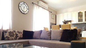 A modern living room with a glass coffee table, a sofa with patterned cushions, a wall clock, and a kitchen area in the background. - Spacious Apartment Neda (Pakostane)