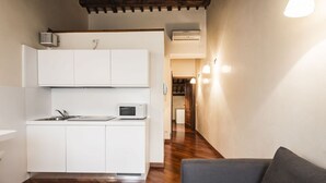 Une cuisine compacte avec des meubles blancs, un four à micro-ondes et un évier. On aperçoit un canapé gris au premier plan et un plancher en bois.