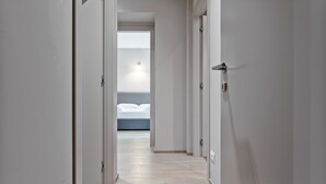 Um corredor estreito com paredes brancas e chão de madeira que leva a um quarto com cama e mesa de cabeceira.