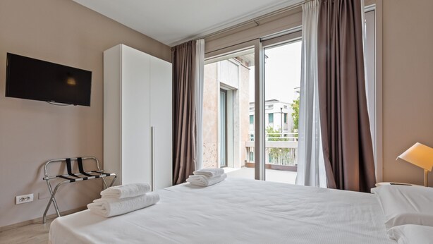 Quarto de hotel com cama, televisão fixada na parede, varanda com cortinas e um porta-toalhas.