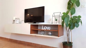 A modern living room with a flat-screen TV mounted on a white wall, a wooden shelf with decorative items, and a potted plant.