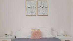 A neatly made bed with a grey bedspread, two pillows, and a rolled towel. There are two bedside tables with lamps and a small plant. The walls are white, and there are two framed pieces of art above the bed.