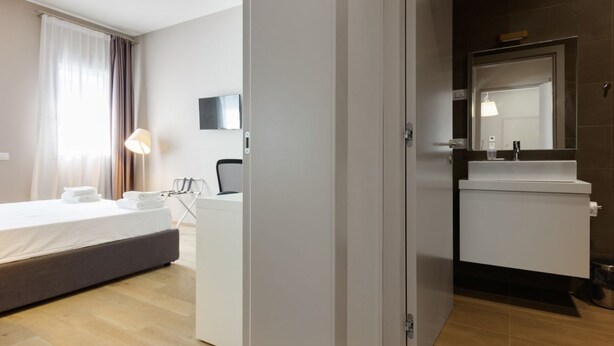 A hotel room with a bed, bedside table, lamp, and a bathroom with a sink and mirror. - MyPlace - Appartamento Family Deluxe Terrace (Treviso)