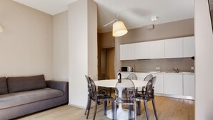 A modern living room with a grey sofa, a round dining table with transparent chairs, and a kitchen area with white cabinets and a microwave. - MyPlace - Appartamento Family Deluxe Terrace (Treviso)