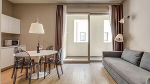 Een moderne woonkamer met een grijze bank, een eettafel met stoelen, een lamp en een schuifdeur van glas die leidt naar een balkon.
